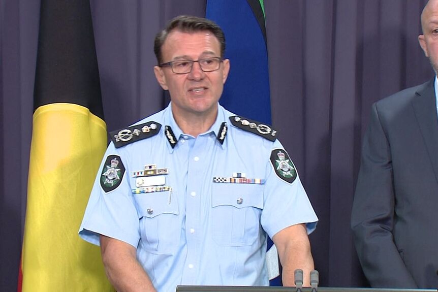 A police officer speaks during a media conference. A man in a suit stands next to him.