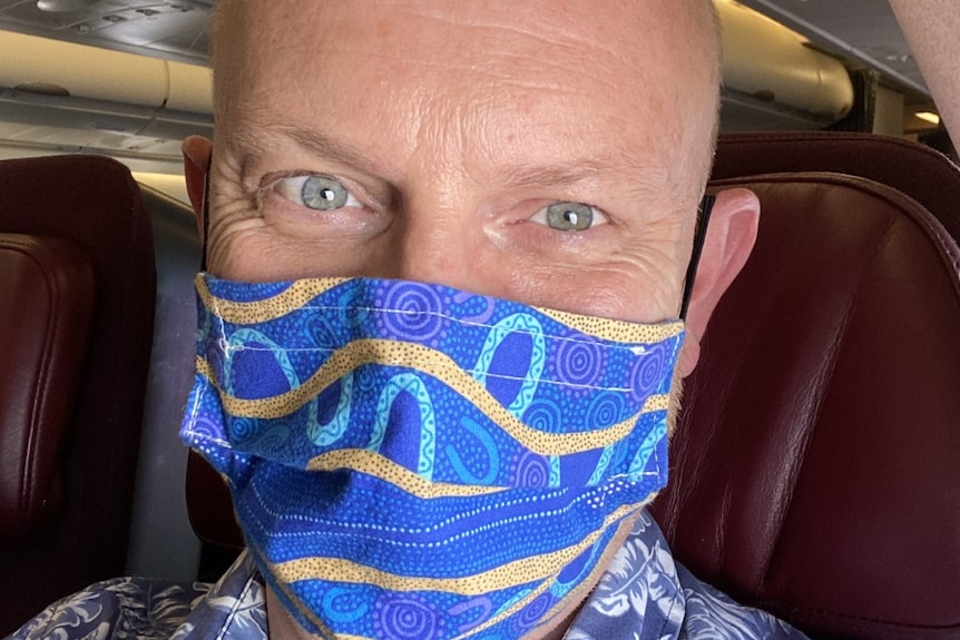 A man smiling underneath his face mask, sitting in an airline passenger seat.