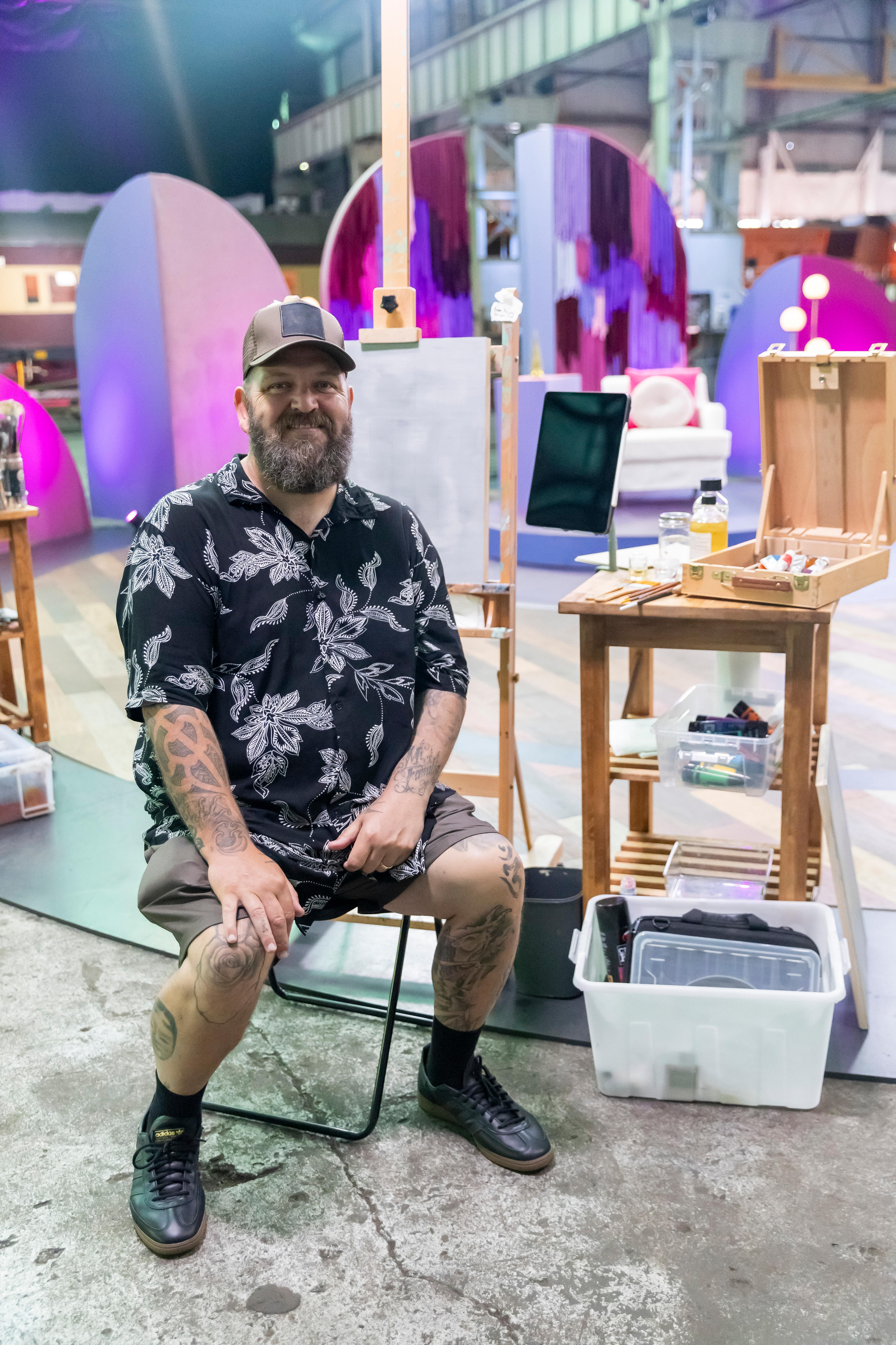 A man with a cap and beard sits next to a table with art supplies on it. He smiles at the camera