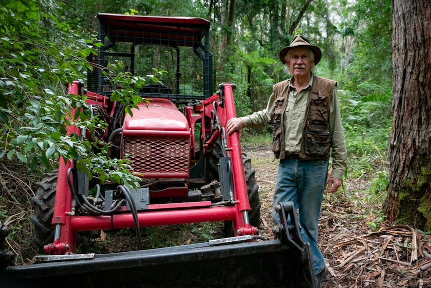 A man stands next to his red tractor on a fire trail he is clearing in a forest.