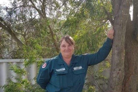 A smiling woman in paramedic's coveralls leaning against a tree.