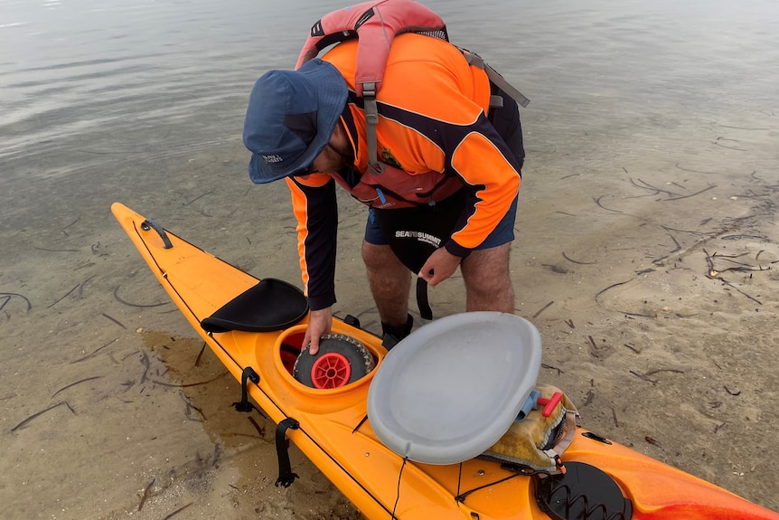 A man wearing high vis bends over a bright orange kayak which is sitting at the edge of a body of water