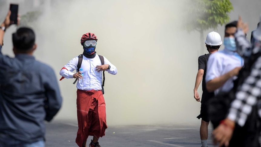 A man in a gas mask wearing Asian sarong runs on street in haze on tear gas within a chaotic crowd.