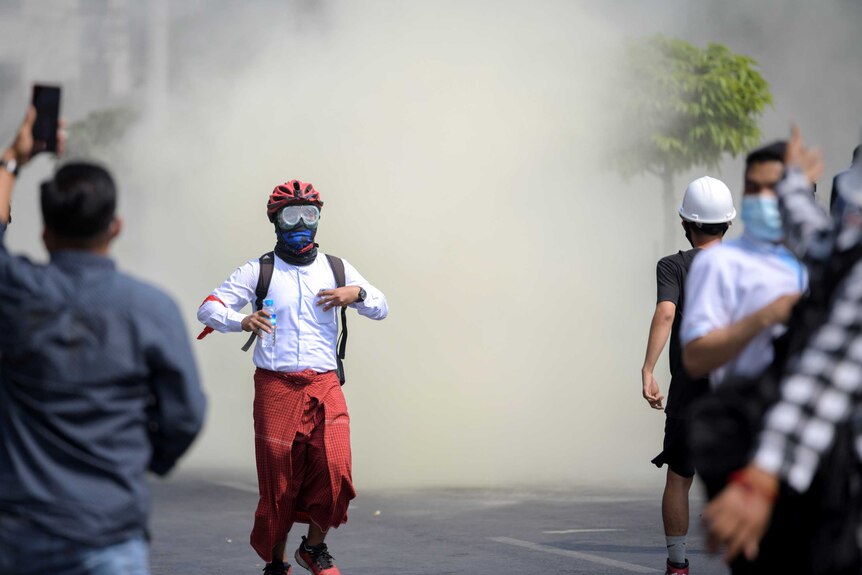A man in a gas mask wearing Asian sarong runs on street in haze on tear gas within a chaotic crowd.