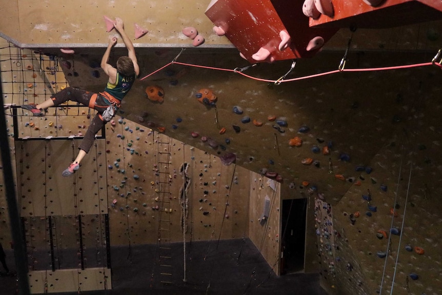 Rock climber Ned Middlehurst clutches onto a training wall as his legs dangle over a drop of several metres.