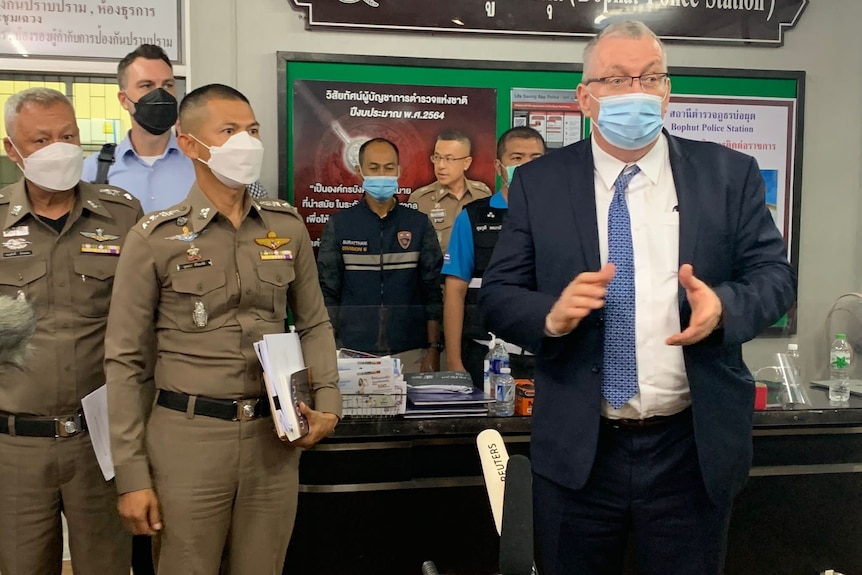 An elderly western man in white hair, face mask and suit speaks as masked Thai police listen on at police station 