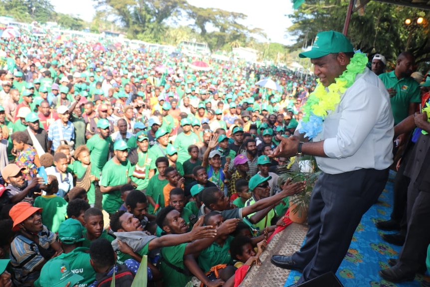 James Marape in a bright green lei around his neck stands on a stage looking out at a huge crowd of people dressed in green