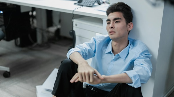 Despondent-looking man in blue shirt sits on ground next to desk.