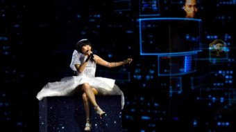 Dami Im wears a white dress as she sits on a cube lit up with stars against an animated dark backdrop while singing.