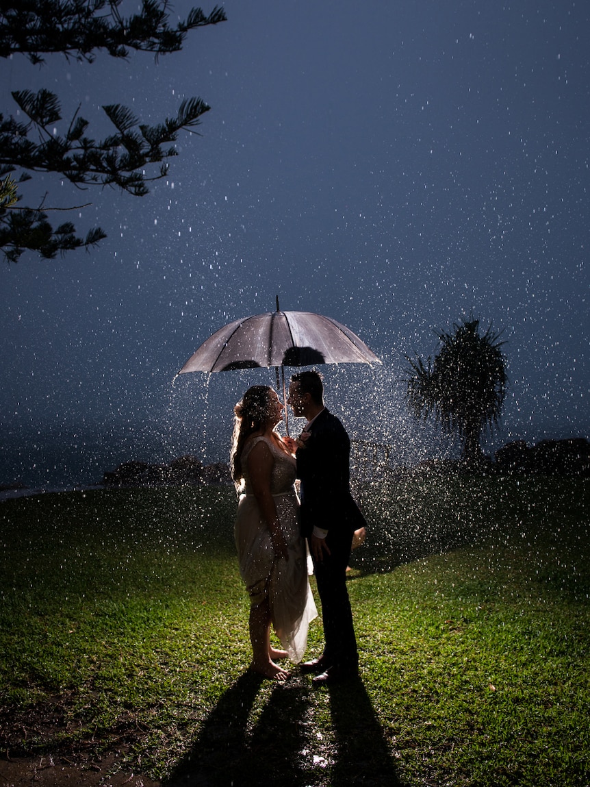 A bride and groom stand under an umbrella as it pours with rain