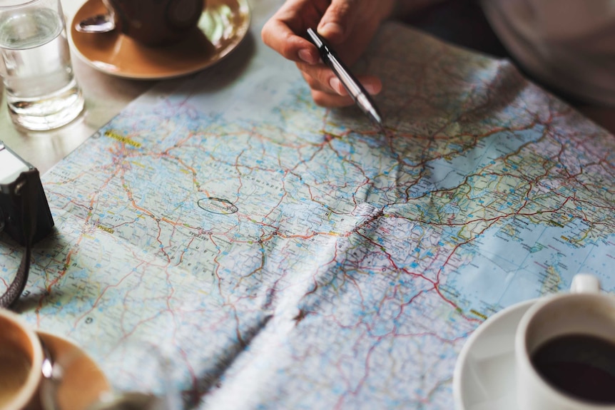 A hand holds a pen above a paper map spread out on a table, weighted down with cups of coffee.