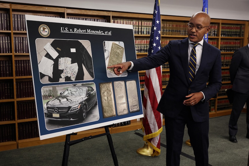 Damian Williams points at a poster board displaying pictures of cash, a convertible and several gold bars.