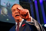 Nigel Farage is shown close up on stage with half of his face obscured by his left hand pointing.