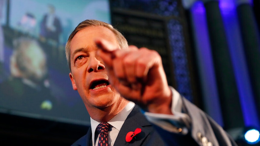 Nigel Farage is shown close up on stage with half of his face obscured by his left hand pointing.