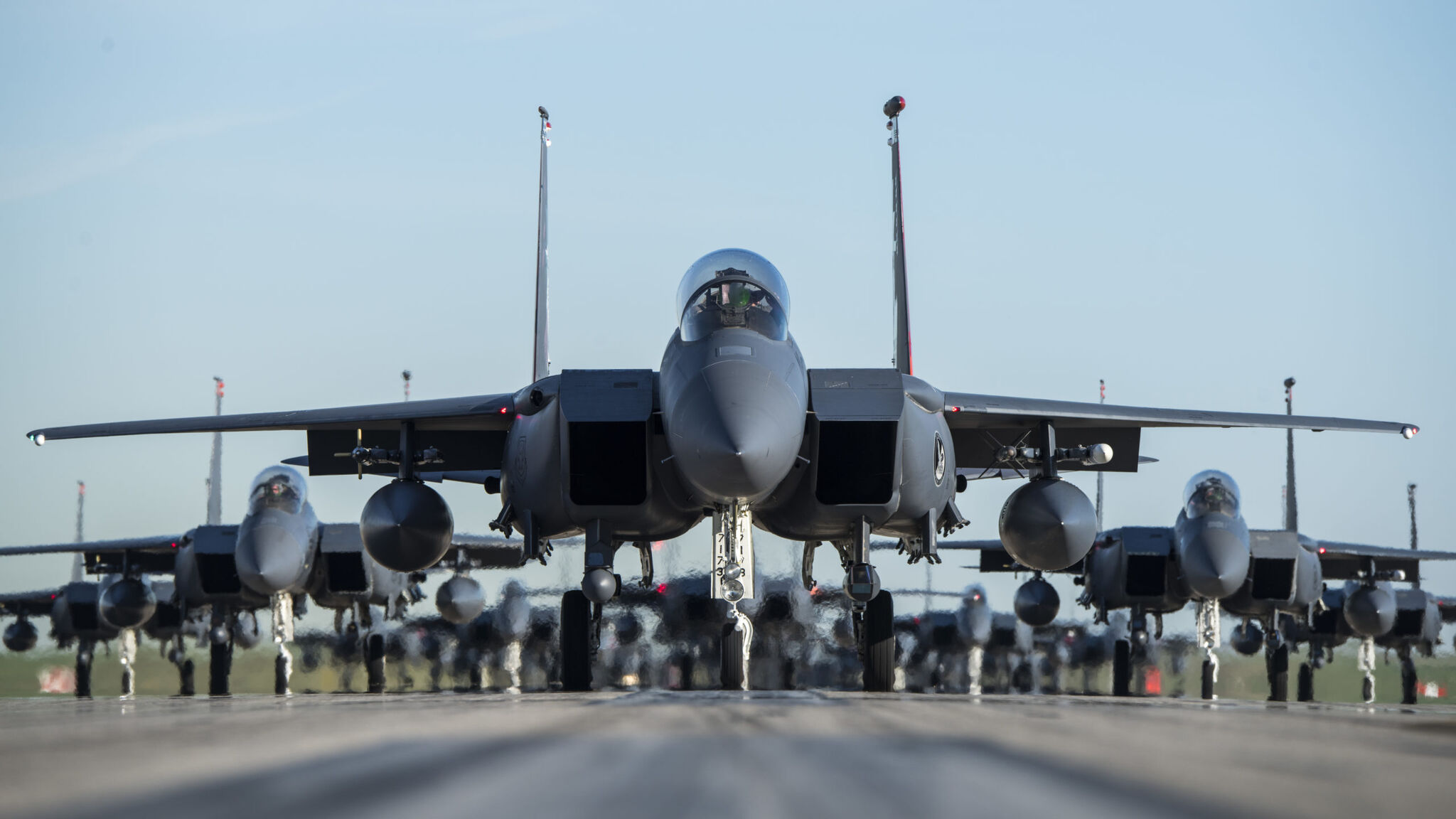 Fighter jets on a tarmac in a formation with one in front with rows splayed behind.