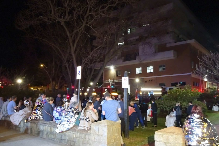 Patients and staff huddle outside a hospital at night with fire trucks in the background.