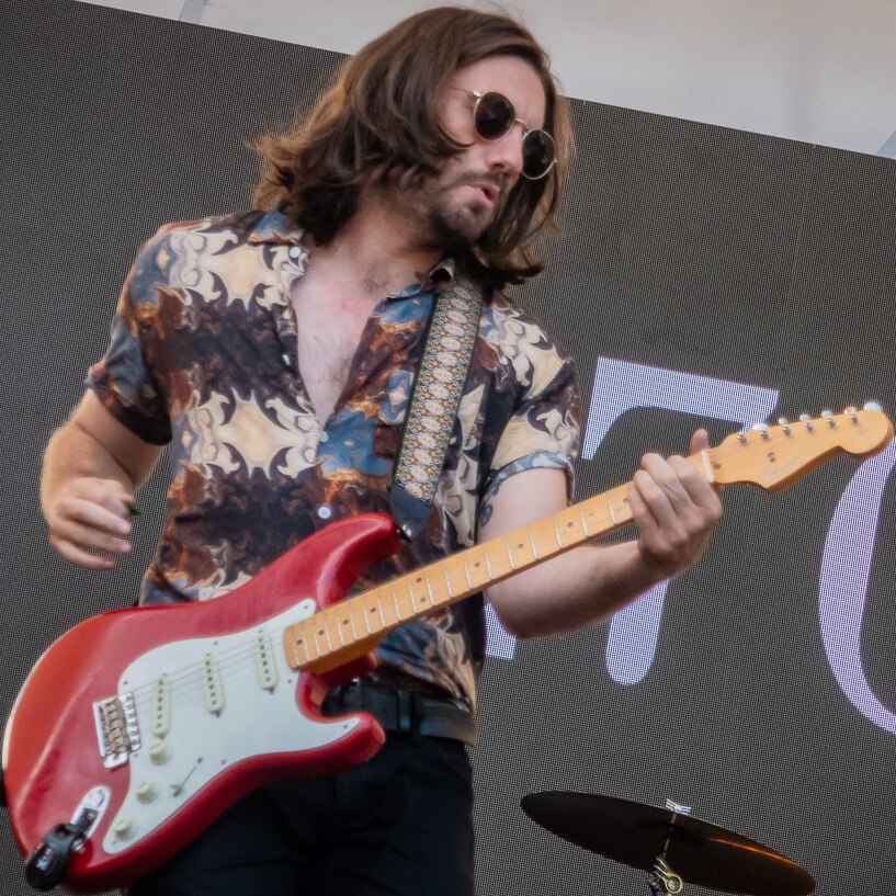 man with long hair and sunglasses on stage in a colourful patterned shirt playing a red guitar