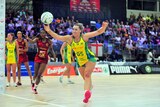 A netball player wearing yellow and green reaches for the ball during a match against a team in red