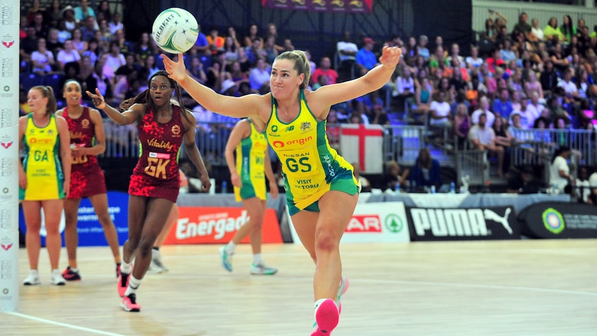 A netball player wearing yellow and green reaches for the ball during a match against a team in red