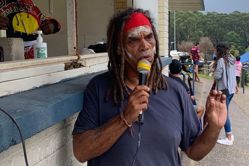 Indigenous man with long dark hair, headband, hold microphone while speaking