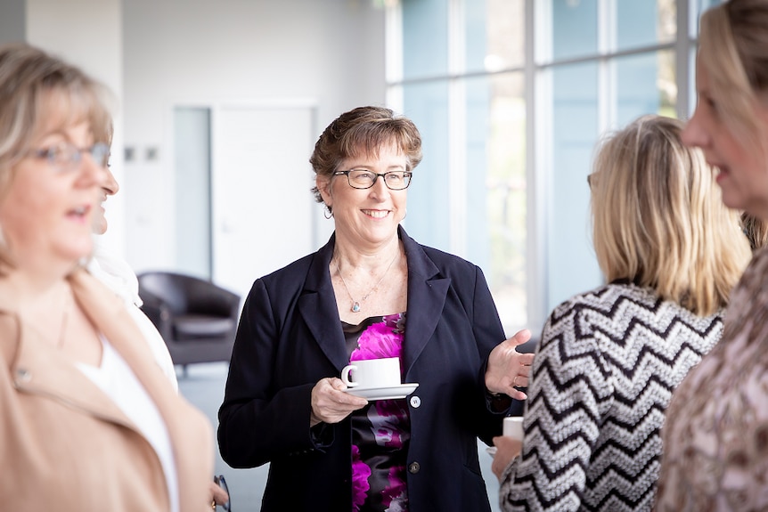 A woman is smiling at another woman talking and holding a cup and saucer. She is wearing a blue blazer and purple and black top.