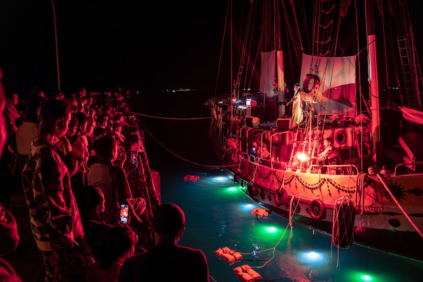Musicians perform on a boat at night in Sydney Harbour in front of an audience, with red lighting illuminating the audience.