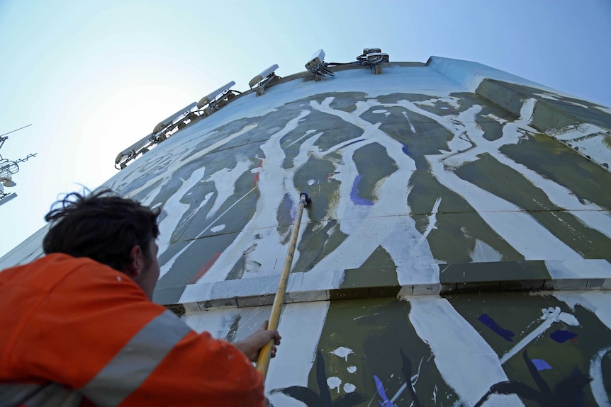 Man a holding a long handle brush looking up the partly painted water tower