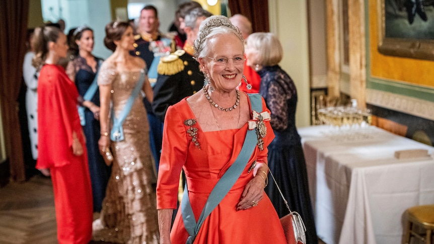 The Queen smiles towards the camera while wearing a red dress with a blue sash. 