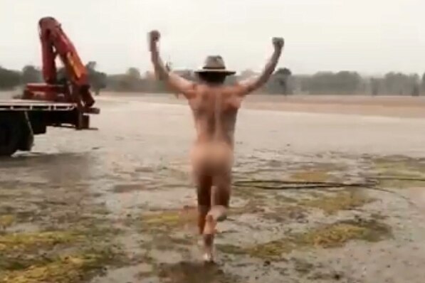A naked farmer wearing only his Akubra hat runs into a rain-soaked field with his fists pumped in the air near Dubbo.