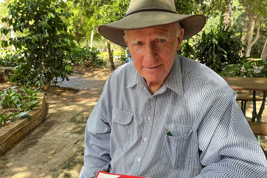 Tom Wyatt, wearing his signature akubra hat, sits at tables at the gardens with lots of trees in the background