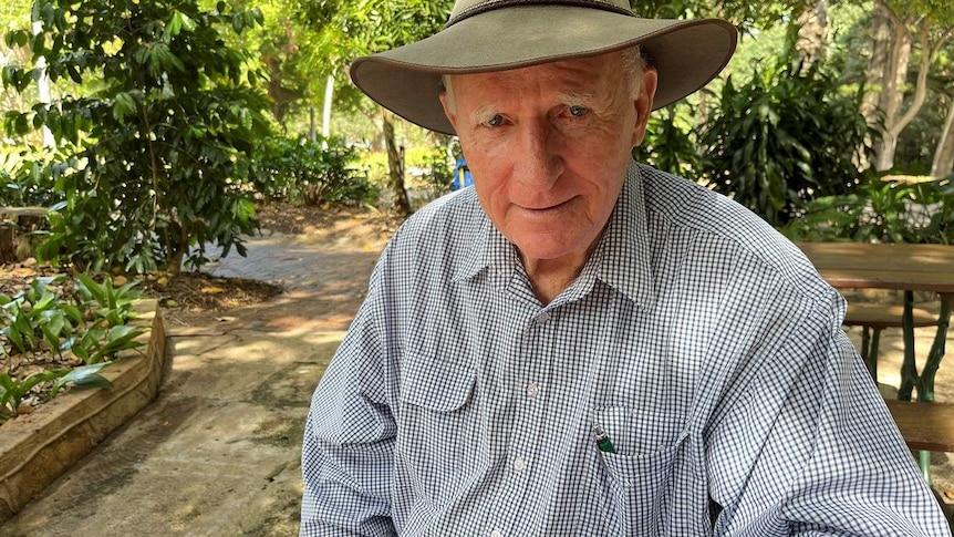Tom Wyatt, wearing his signature akubra hat, sits at tables at the gardens with lots of trees in the background