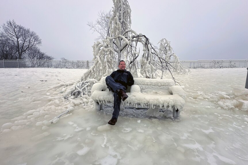 A man sits on a frozen bench in front of an ice-covered tree, the ground is frozen