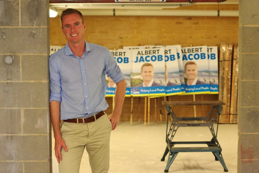 Albert Jacob leans against a concrete wall with campaign signs in the background.
