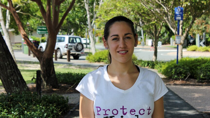 A young woman wearing a 'protect kids' t-shirt stands on a shaded footpath.