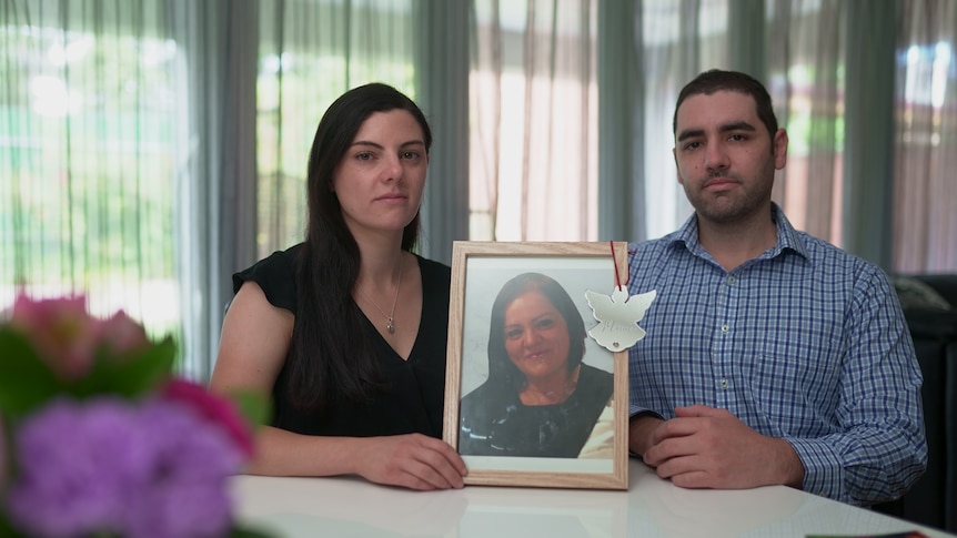 A woman and man seated at a table, holding a framed photo of their mum. Their expressions are sombre