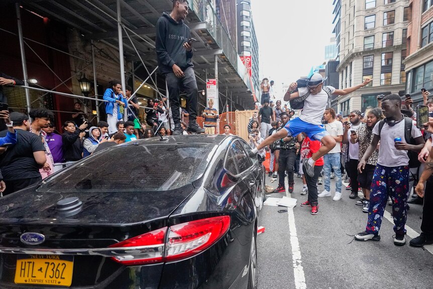One young man stands on top of a car and another jumps up to kick the car's window as a crowd watches on