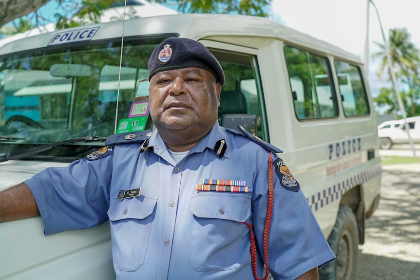A man in a police uniform and beret leans against a van with palm trees in the background