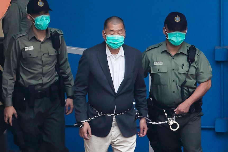 A man wearing a suit and mask is escorted in chains by two uniformed police wearing black caps and masks