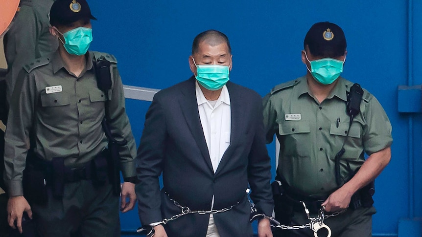 A man wearing a suit and mask is escorted in chains by two uniformed police wearing black caps and masks