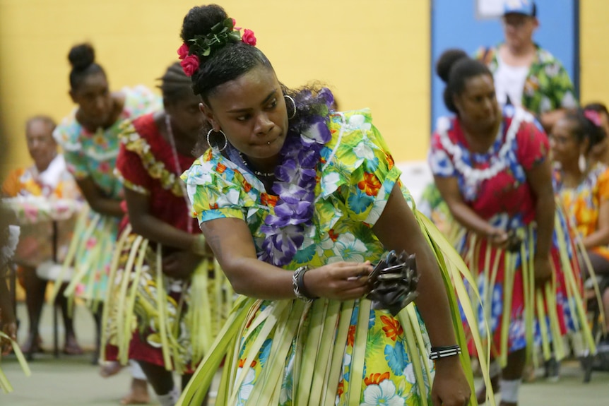 Woman in floral dress with lei dances with seeded rattle