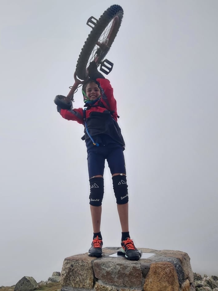 An 11-year-old boy in bicycle wear holds a unicycle over his head, at the top of a mountain.