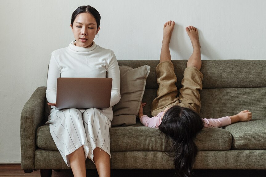Woman sitting on a couch working with laptop while child puts feet on the wall  