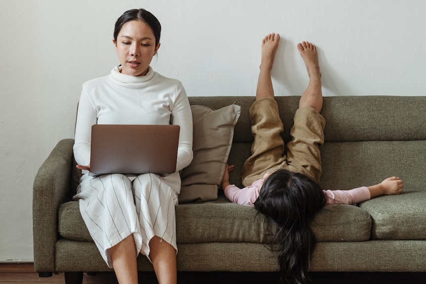 Woman sitting on a couch working with laptop while child puts feet on the wall  