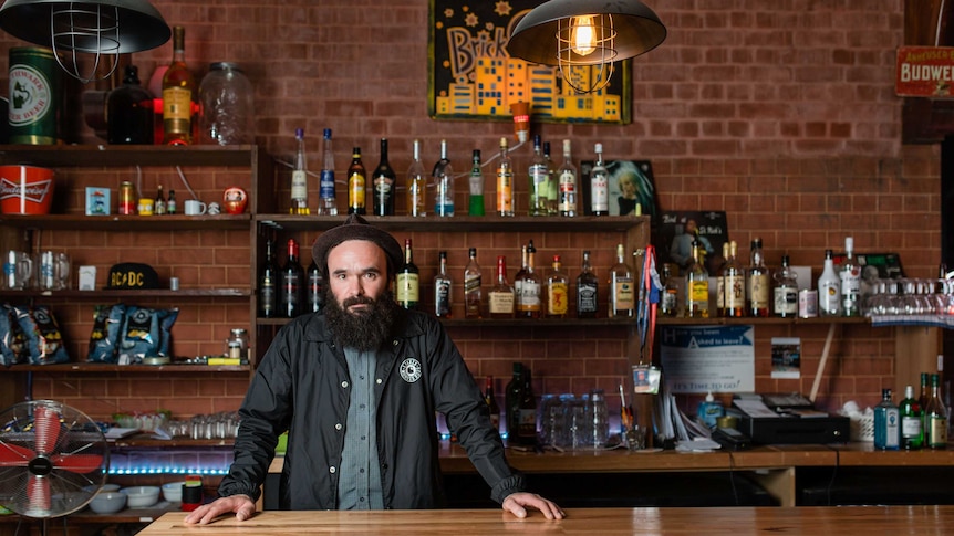 Bearded man Steve Faragi standing in a bar, surrounded by liquor, looking determined.