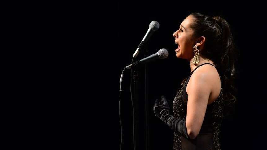 A profile photo of jazz vocalist Veronica Swift singing into a microphone in a black evening dress and elbow length gloves.