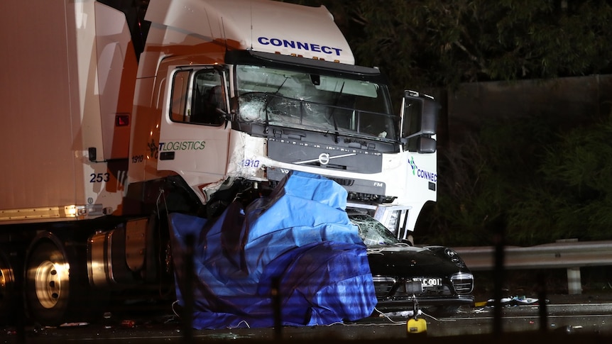 The wreckage of a black car is crushed underneath a large refrigerated truck in an image taken at night.