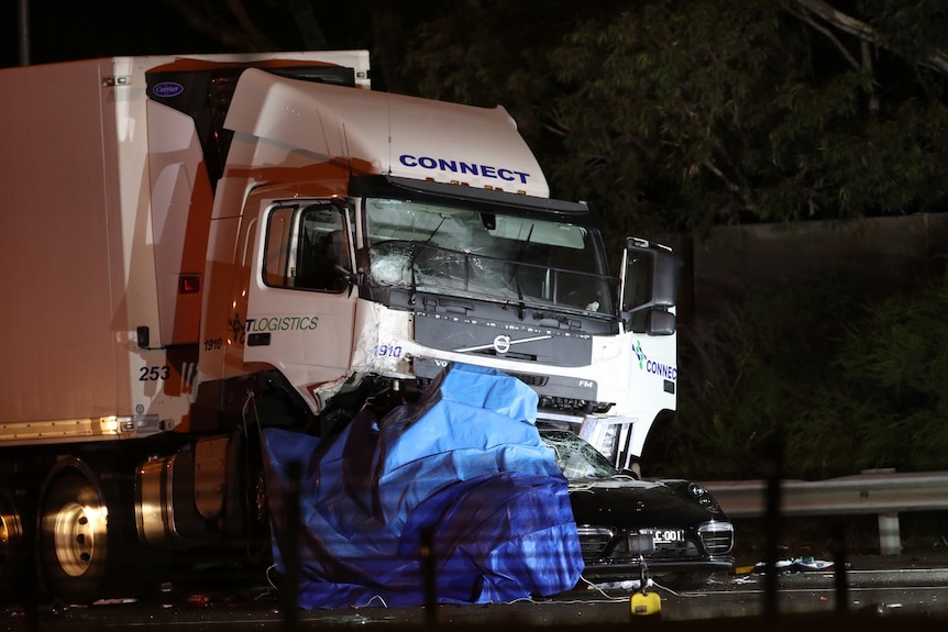 The wreckage of a black car is crushed underneath a large refrigerated truck in an image taken at night.