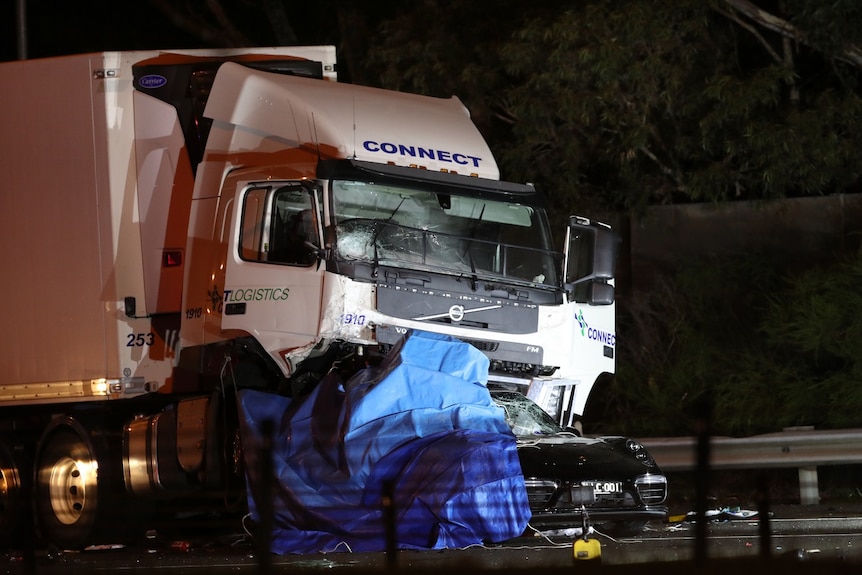 L'épave d'une voiture noire est écrasée sous un gros camion frigorifique sur une image prise de nuit.