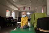 A child with a bald head sits in an empty hospital hallway with a mask on.