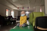 A child with a bald head sits in an empty hospital hallway with a mask on.